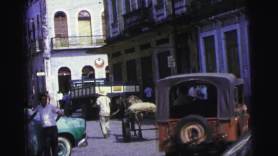 The horse cart and red Jeep-style vehicle share a cobblestone street in 1967 Brazil, with a green car and pedestrians at the edge.