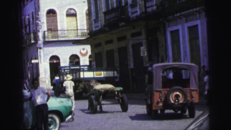 A red Jeep-style vehicle recedes beside a horse-drawn cart on a 1967 Brazil street lined with arched buildings and pedestrians.