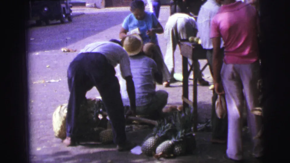 1967 Brazil 8mm frame showing people around a pineapple pile, including a seated man in a straw hat and others standing nearby.