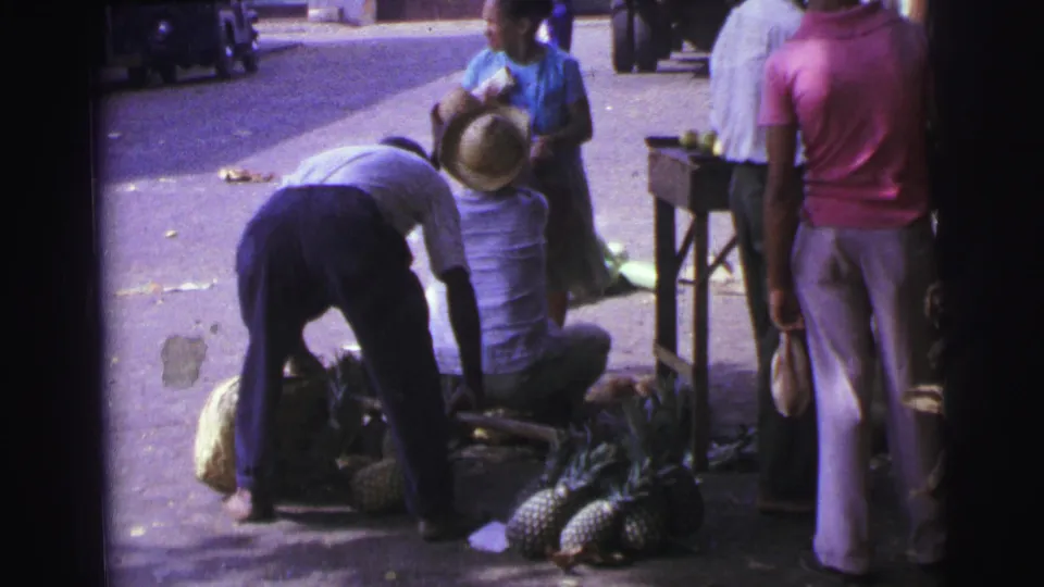 1967 Brazil market frame with a person bending near pineapples, a seated man in a straw hat, and shoppers beside a small stall.