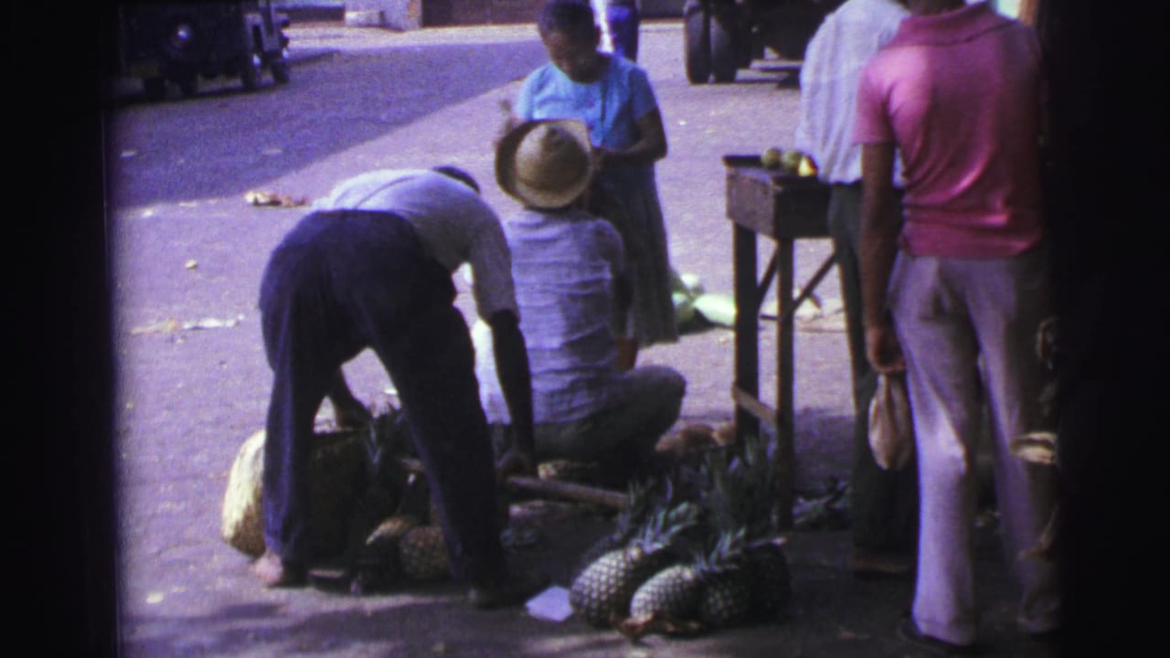 Men gathered on a sidewalk beside pineapples and other produce.
