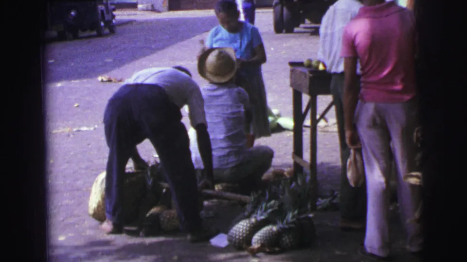 1967 Brazil home-movie frame showing men on a sidewalk around pineapples and produce, with one man wearing a straw hat.