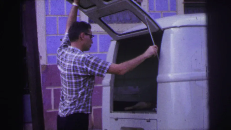 1967 Brazil frame of a man in glasses and a plaid shirt holding open the rear compartment of a white van near a tiled wall.