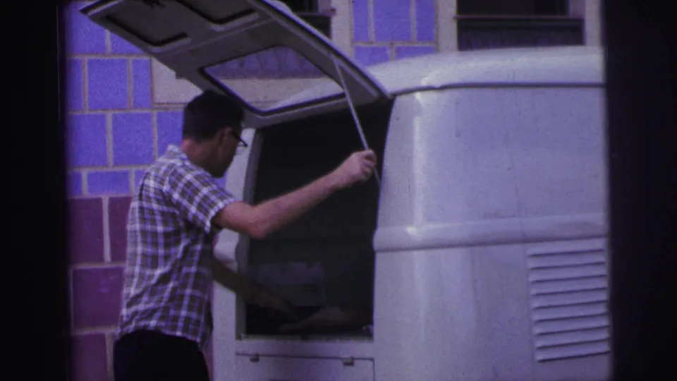1967 Brazil home-movie frame showing a plaid-shirted man working at the open rear of a white van against a blue tiled wall.