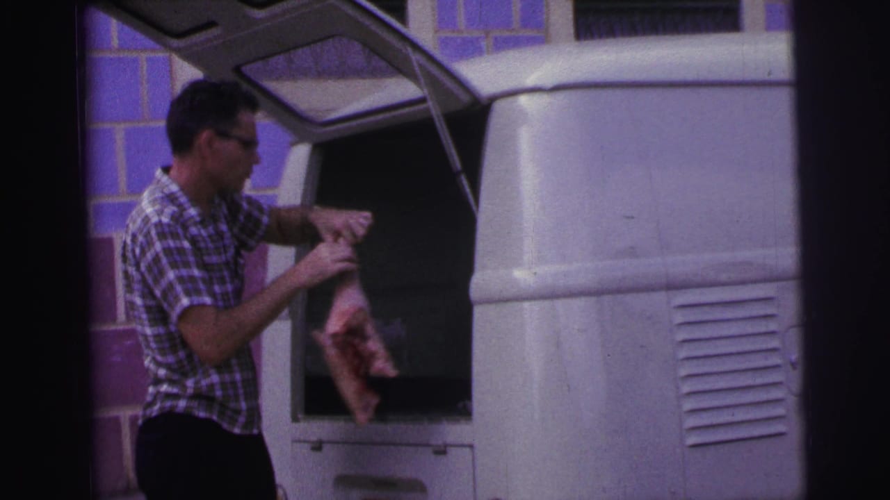 Man in plaid shirt reaches into the open back of a white van beside a purple-tiled wall.