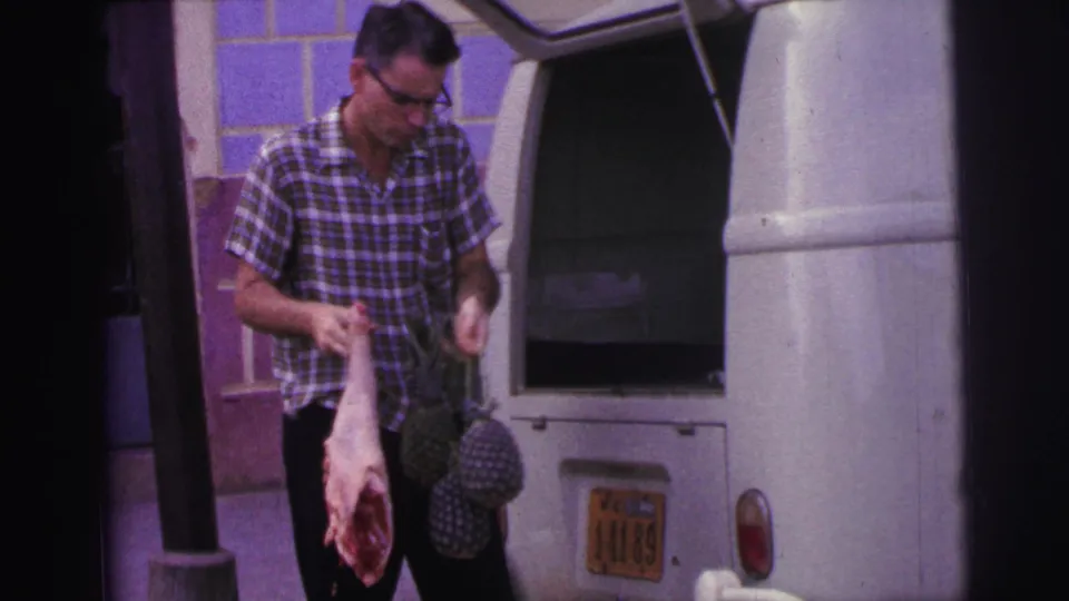 In 1967 Brazil footage, the man holds raw meat and pineapples beside the open white van and tiled exterior wall.