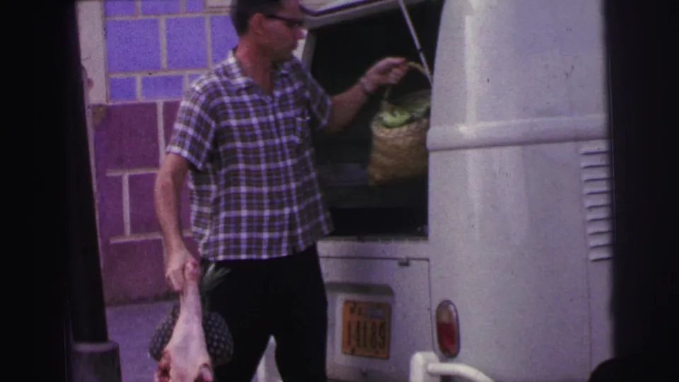 The man reaches into the open van toward sacks of pineapples while still holding raw meat at his side.