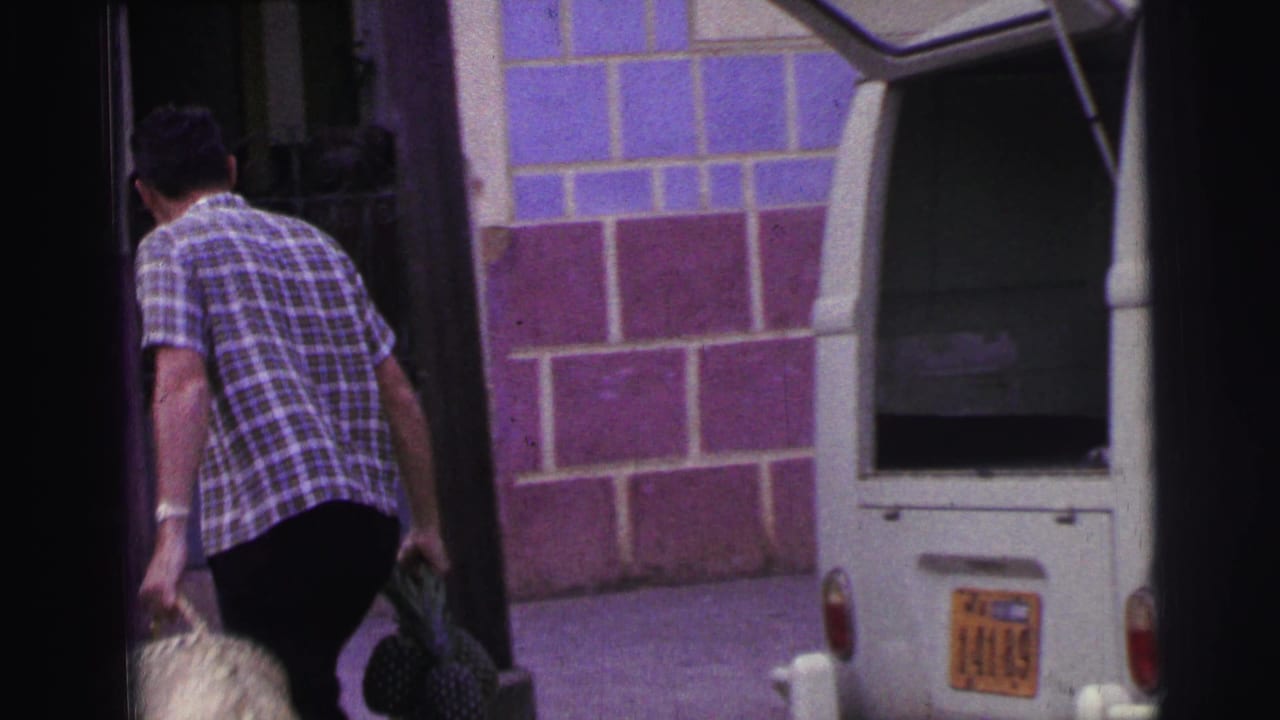 Man in a checkered shirt carries bags beside a tiled wall and a white car.