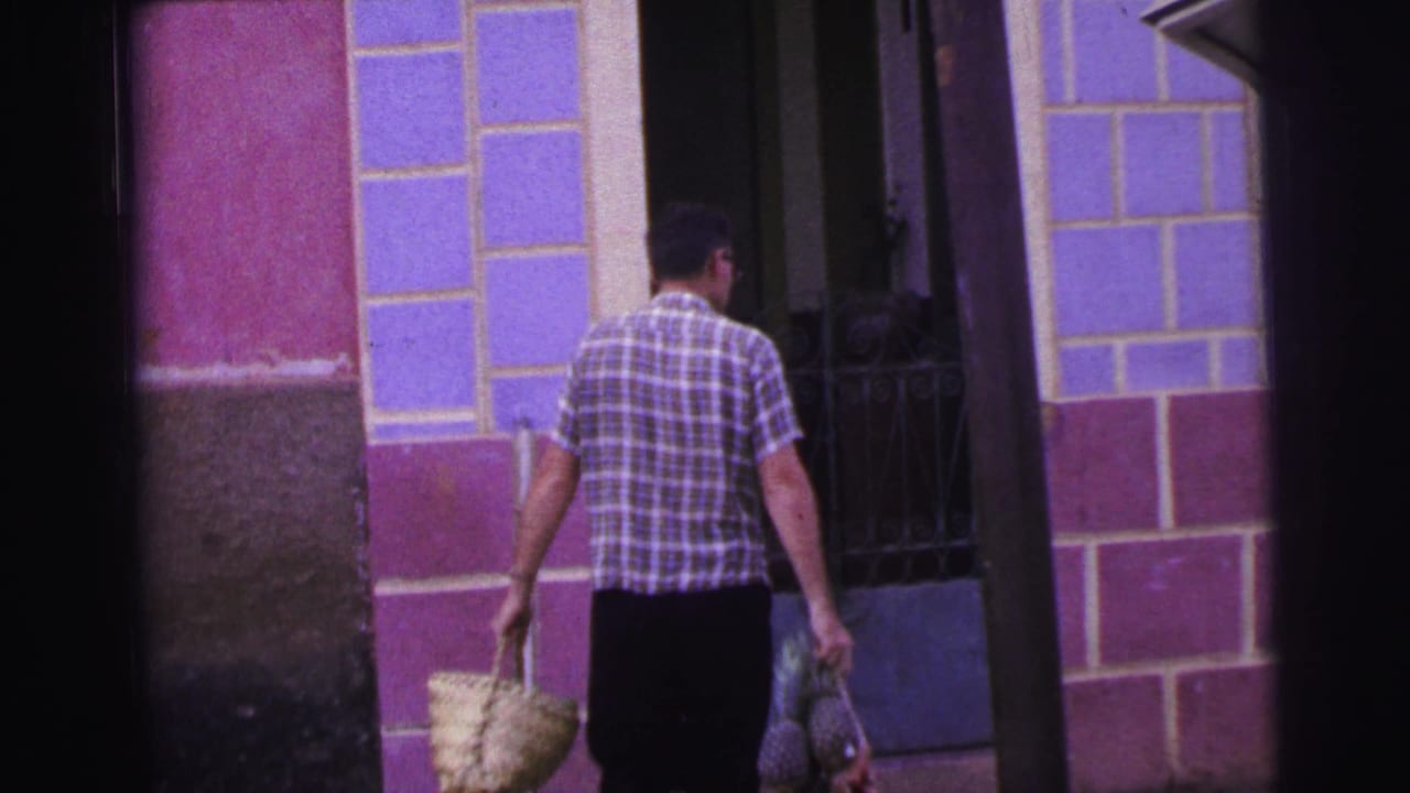 Man walks toward a doorway carrying a basket and bundle against a tiled wall.