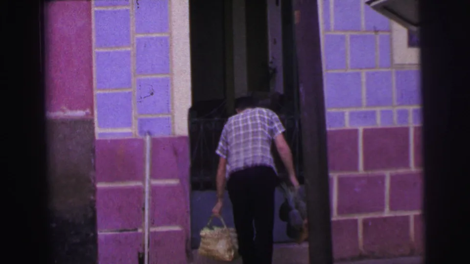 In the home-movie frame, the man bends forward at the doorway with a woven basket in hand and another bundle near the entrance.