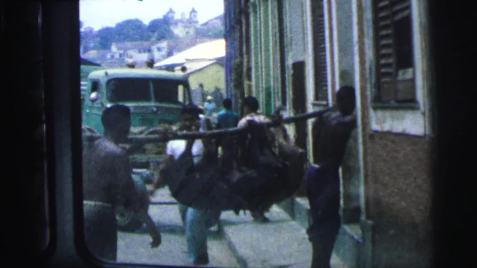 1967 Brazil 8mm frame showing men in a narrow lane carrying a large animal on a pole, with a truck ahead.