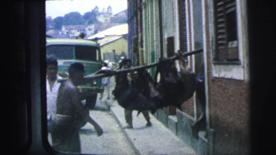 1967 Brazil frame showing several men with a large animal on a pole beside building facades in a narrow alley.