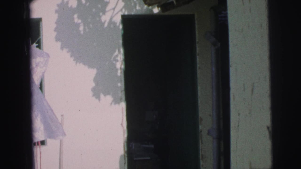 A 1967 Brazil home-movie frame with a dark doorway, pale wall, and a light-colored figure or garment at the entrance.