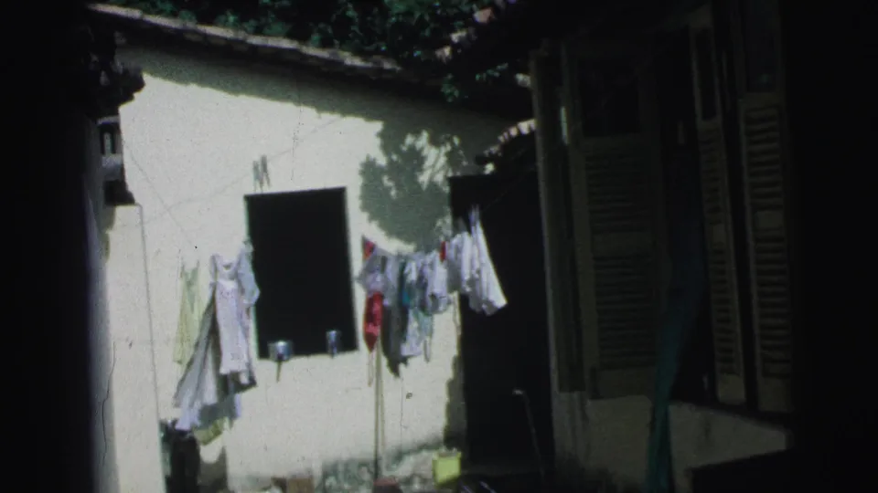 1967 Brazil home-movie frame showing laundry on a clothesline against a pale courtyard wall with dark shutters nearby.