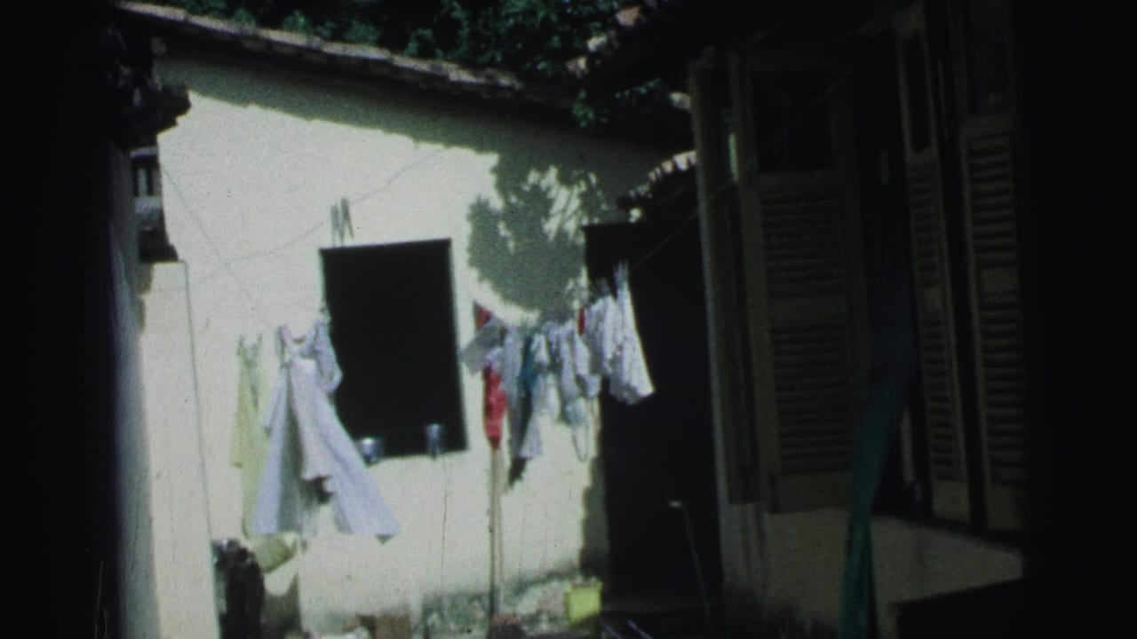 Sunlit courtyard wall with laundry hanging on a clothesline beside dark shutters and a doorway.