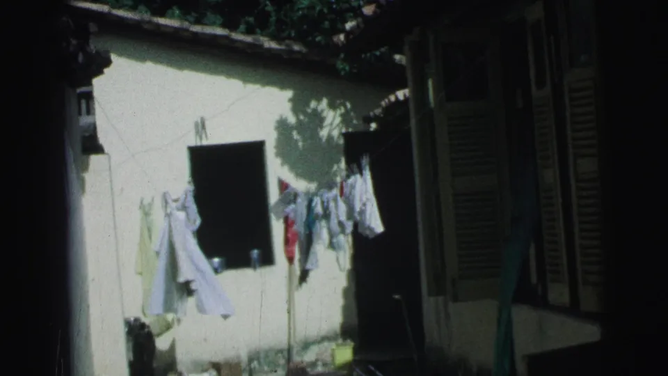 1967 Brazil 8mm frame showing a bright courtyard wall, hanging laundry, louvered shutters, and a shaded doorway.