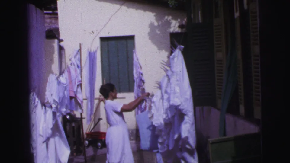 1967 Brazil home-movie frame showing a woman tending white garments on a clothesline beside shutters and a red wagon.