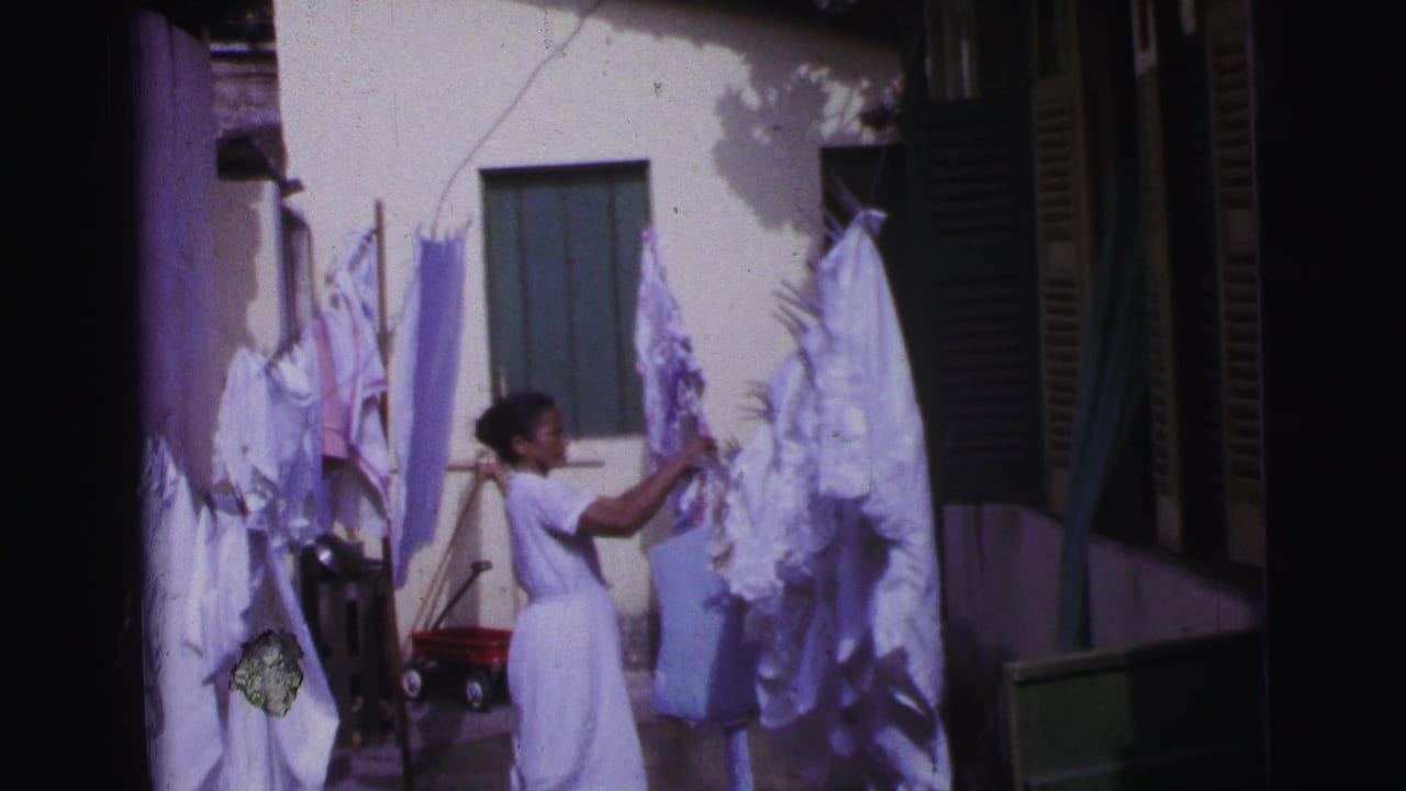 Woman in a white dress reaches toward laundry hanging on a sunlit courtyard clothesline.