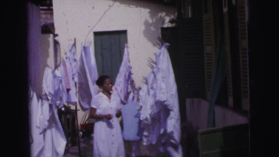 In 1967 Brazil, the woman pauses among white linens on the clothesline, with a cream wall, green shutters, and dark doorway nearby.