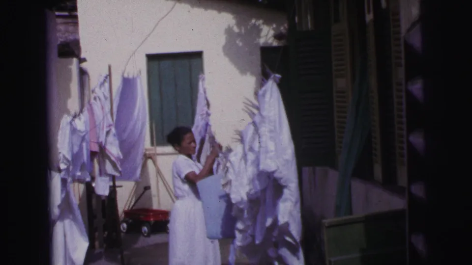 In 1967 Brazil, the woman raises a white garment or linen toward the clothesline as courtyard shadows fall across the wall.