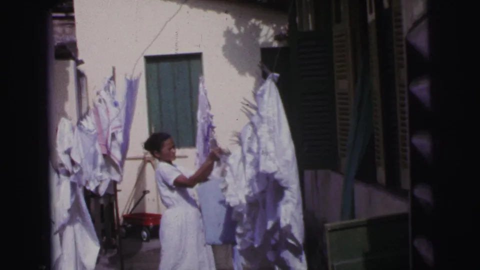 In 1967 Brazil, the woman adjusts white linens on a crowded clothesline in a sunlit courtyard with green shutters and a red wagon visible.