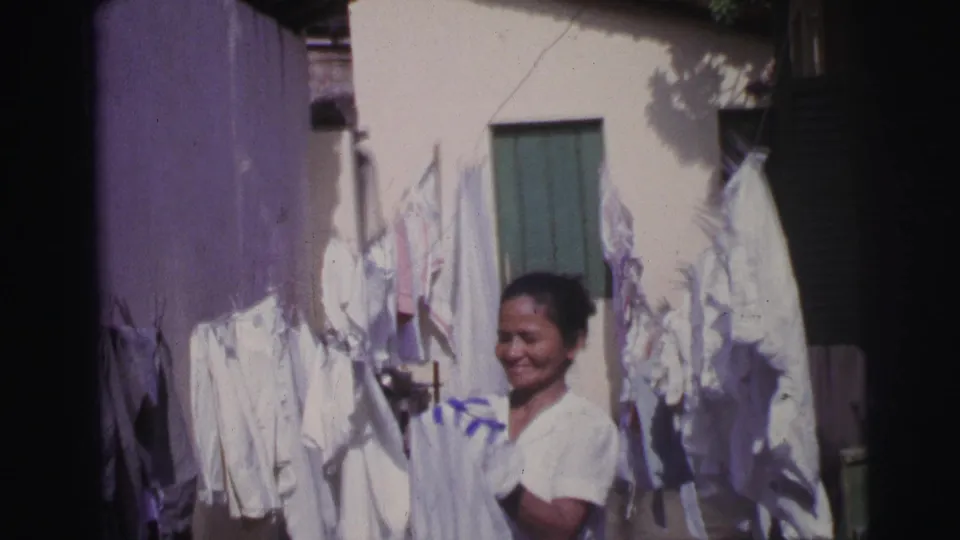 In Brazil, 1967, a smiling woman holds a white garment near clotheslines against a cream building with green shutters.