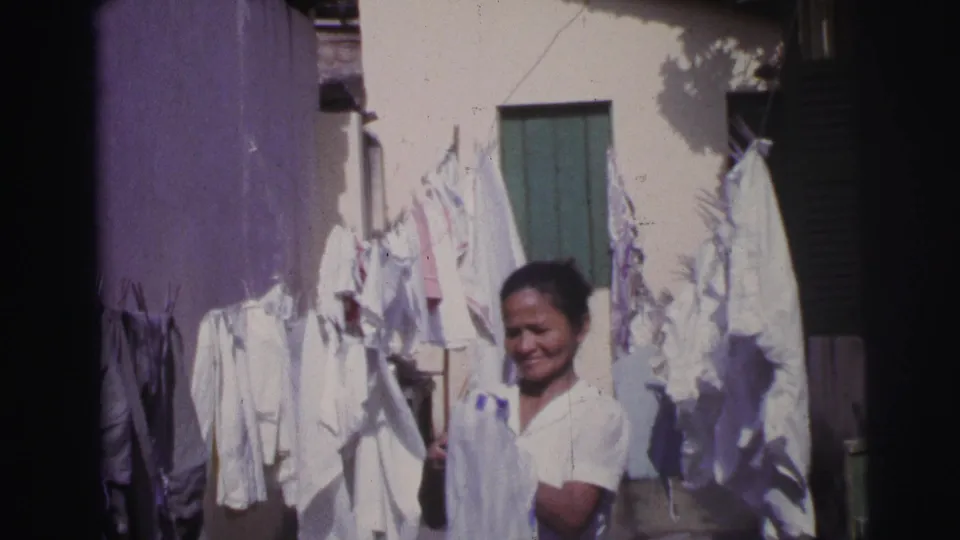 In Brazil, 1967, the woman stands close to hanging white garments, with a green shutter and sunlit wall behind her.