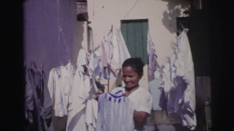 In Brazil, 1967, the woman smiles while holding a white garment under lines of drying clothes in the courtyard.