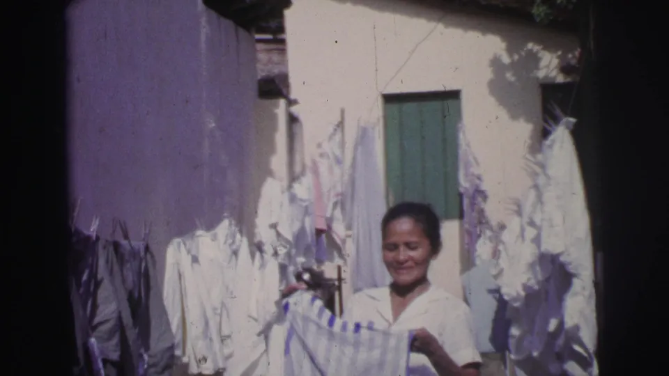 1967 Brazil 8mm frame of a woman folding laundry in a courtyard, surrounded by white garments and sunlit walls.