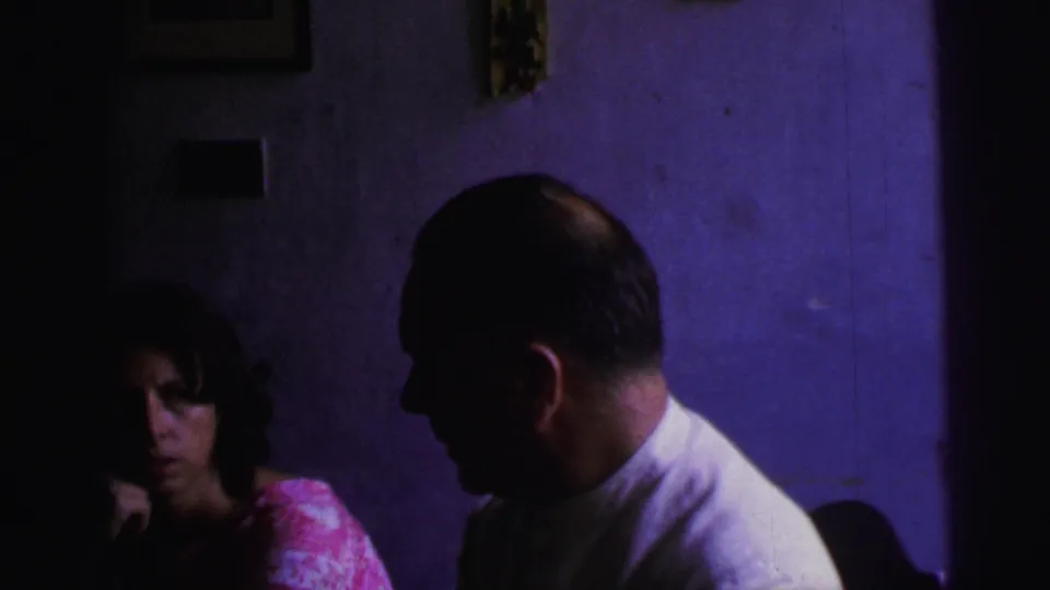 1967 Brazil home-movie frame showing two adults in a dark interior, including a man in profile and a woman seated in shadow.