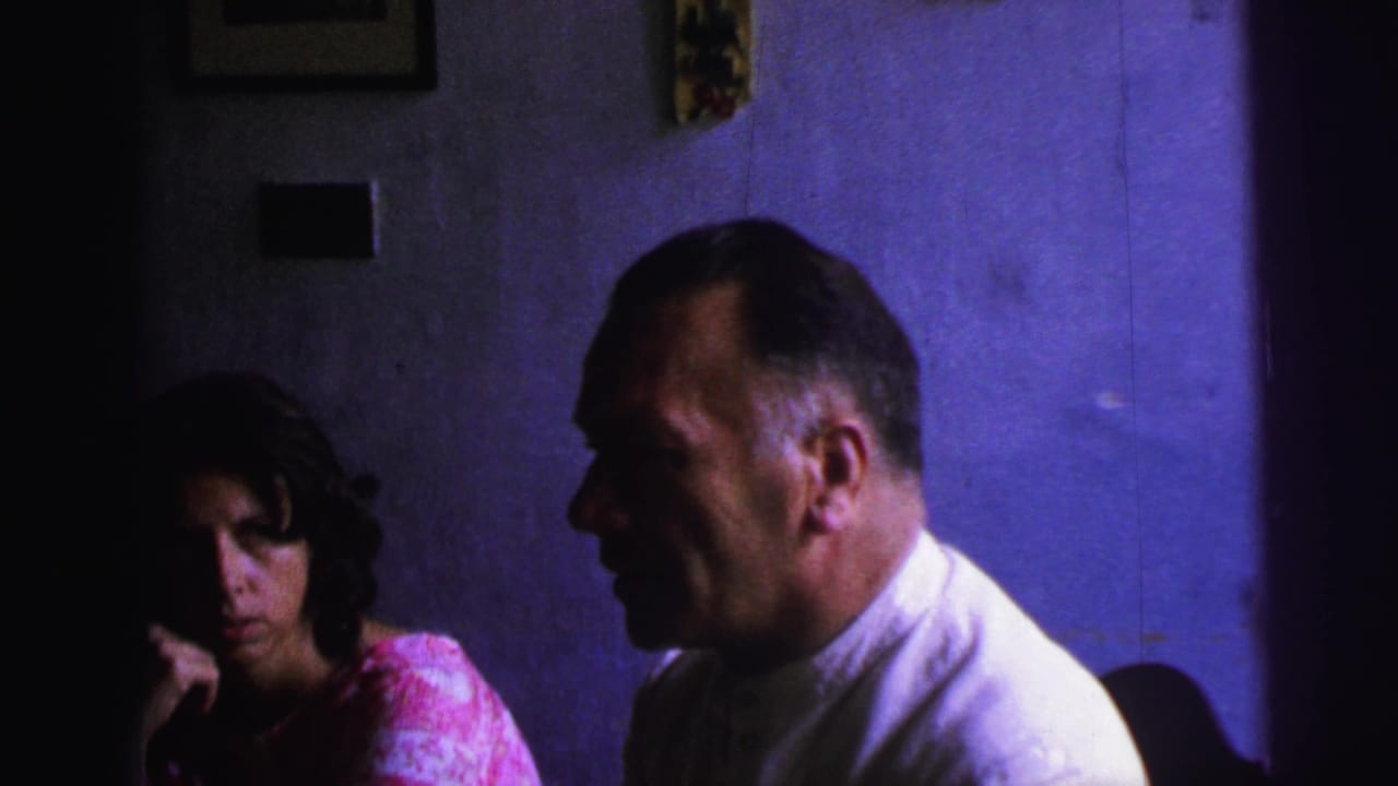 1967 Brazil 8mm frame showing a seated man in a white collared shirt turned toward a woman in a pink patterned dress in a dark interior.