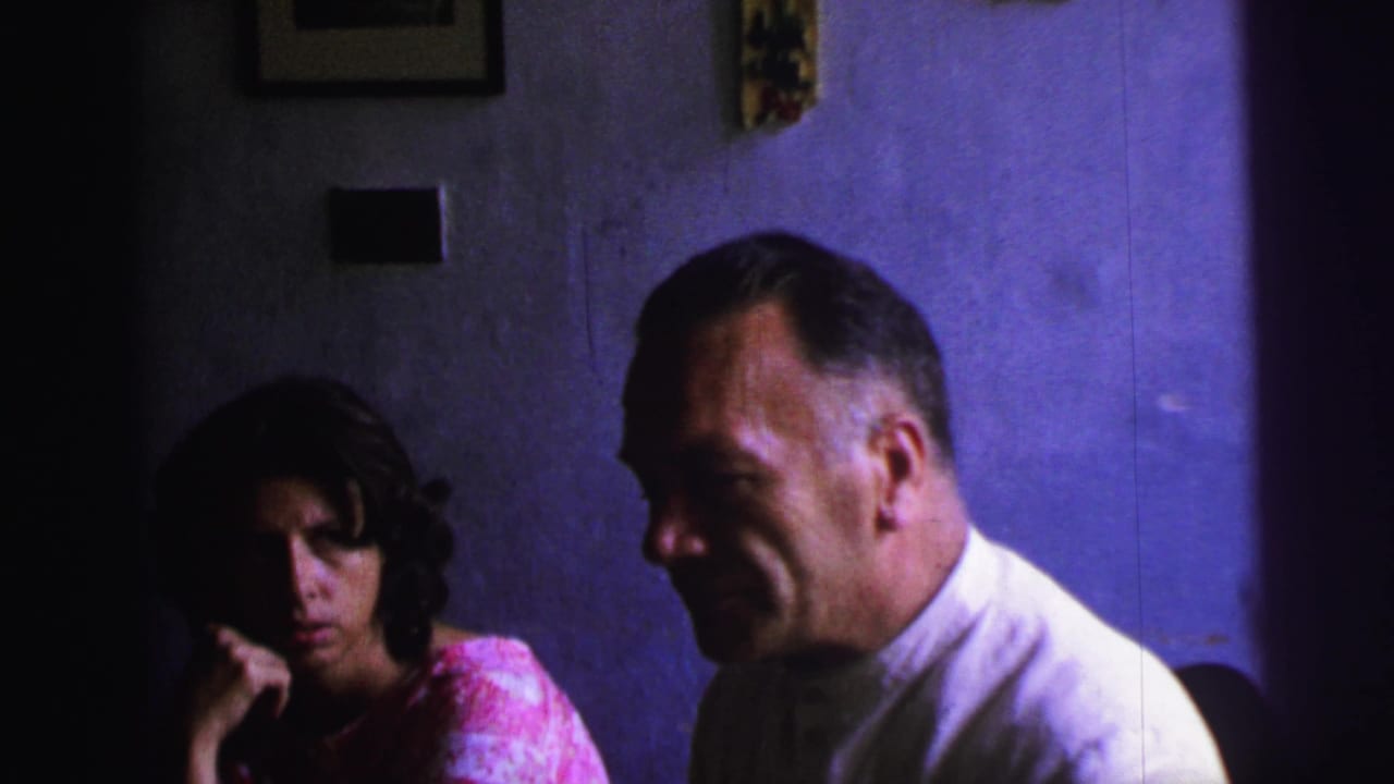 1967 Brazil home-movie frame with a woman listening at left and a formally dressed man seated in profile against a purple wall.