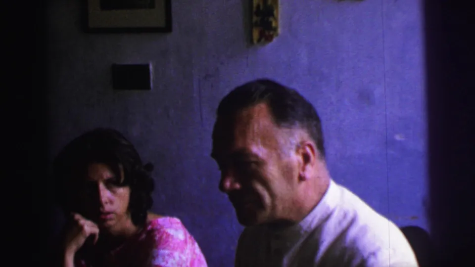 1967 Brazil home-movie frame with a woman listening at left and a formally dressed man seated in profile against a purple wall.