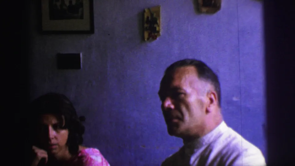 1967 Brazil home-movie frame showing a man in a white shirt and a woman in a pink dress seated in a dim domestic interior.