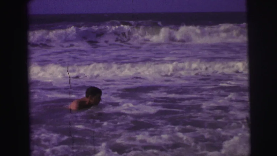 1967 Brazil 8mm frame of a swimmer in profile moving through foamy ocean water with larger waves behind.