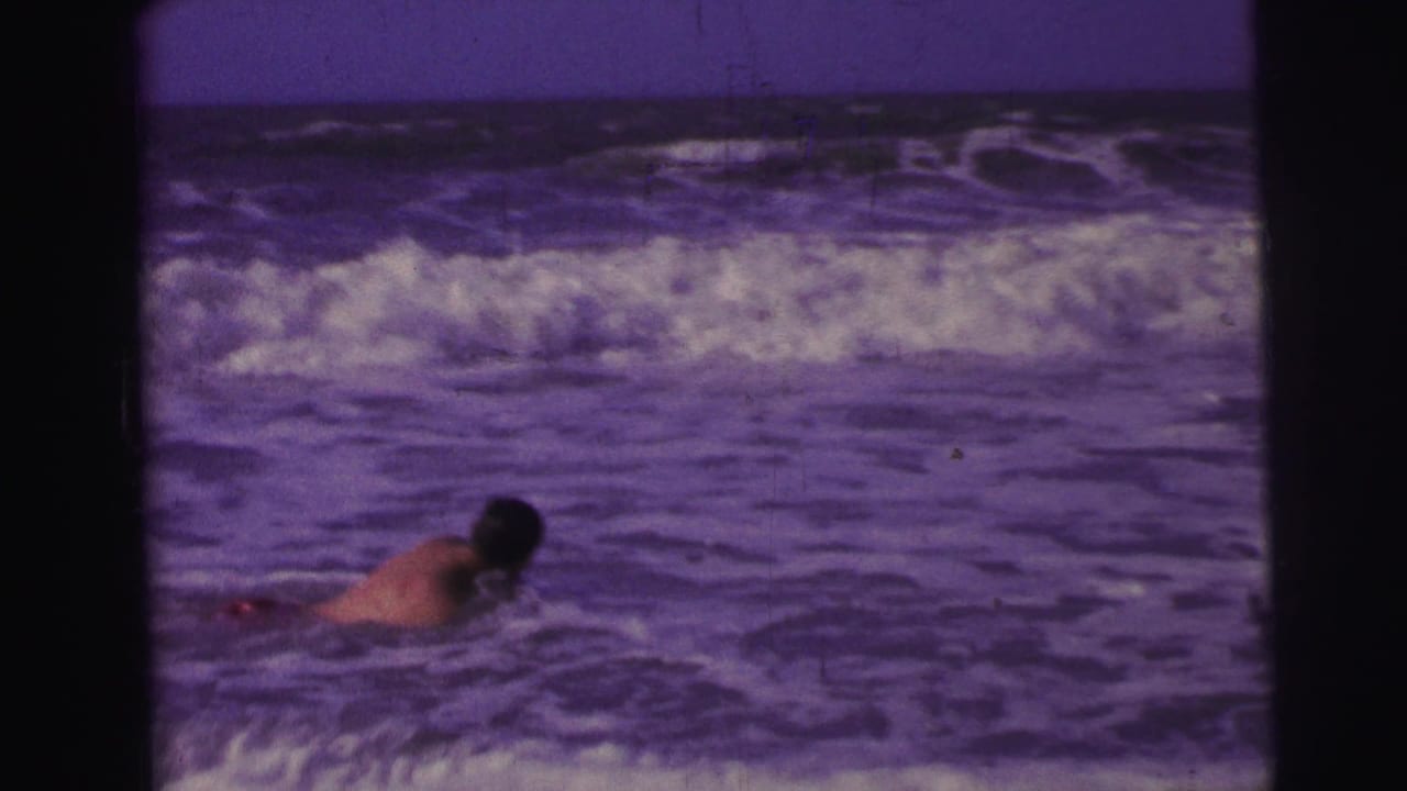 1967 Brazil 8mm frame showing a man in red swim trunks partly submerged as waves break behind him.