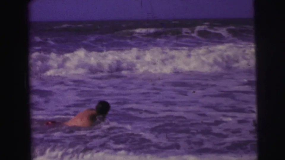 1967 Brazil 8mm frame showing a man in red swim trunks partly submerged as waves break behind him.