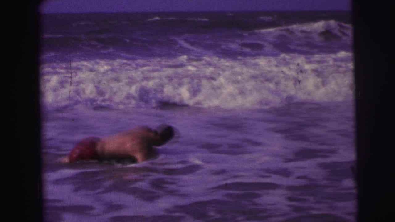 Man lies forward in shallow ocean water with waves breaking behind him.
