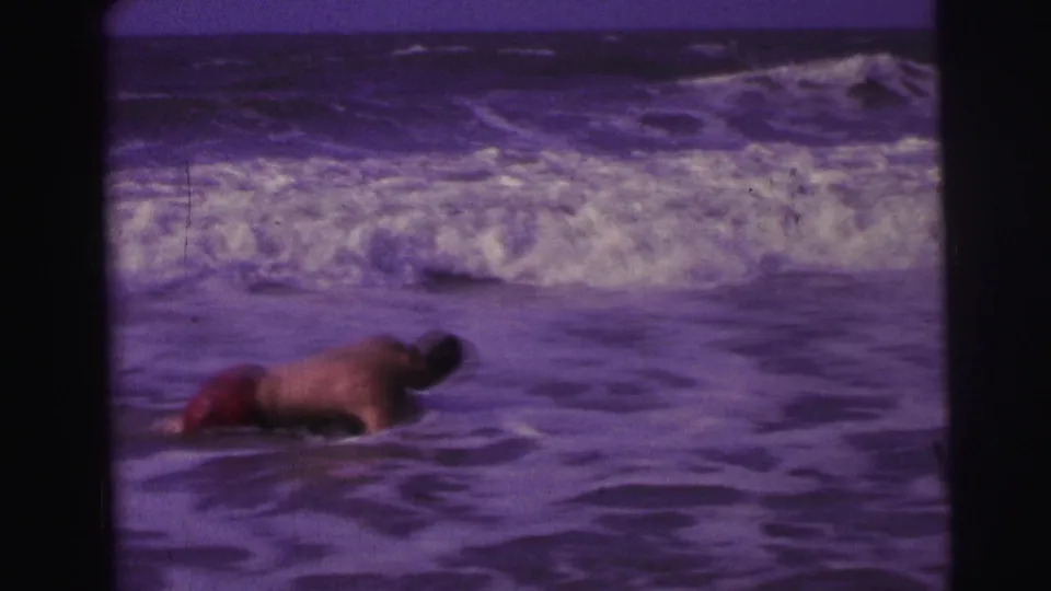 1967 Brazil beach footage frame with a man in red swim trunks leaning forward in the surf under a purple color cast.