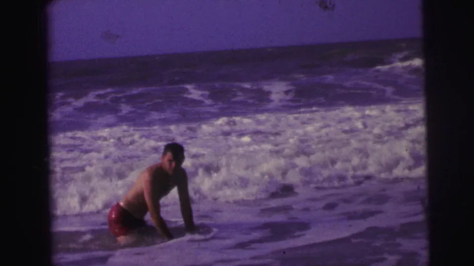 1967 Brazil 8mm frame showing a man in red trunks rising or kneeling in shallow surf with breaking waves behind him.