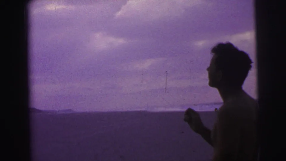 In 1967 Brazil home-movie footage, a silhouetted man stands by the beach beneath a purple-tinted sky.