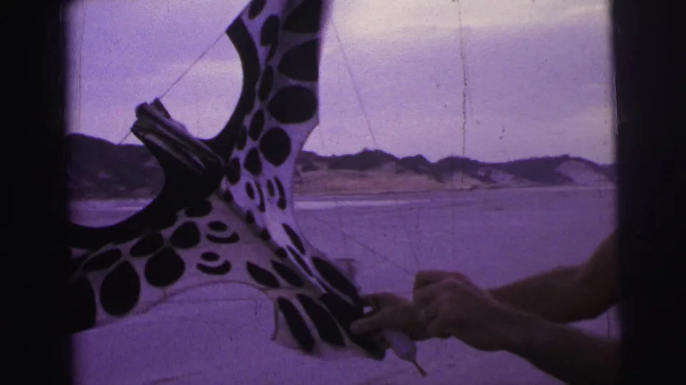 In 1967 Brazil home-movie footage, a shirtless man adjusts a black-and-white spotted kite on a windy beach.