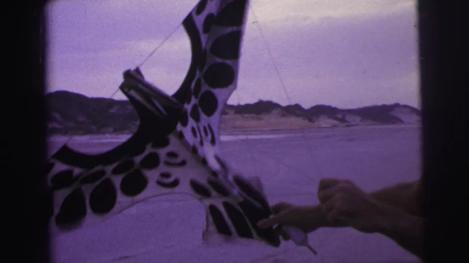 The man’s hands work near the frame of a black-and-white spotted kite on a Brazilian beach in 1967 footage.