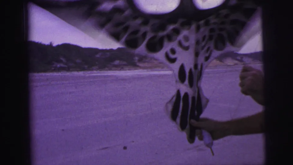 A spotted bird-shaped kite flutters close to the camera over a sandy beach in 1967 Brazil home-movie footage.