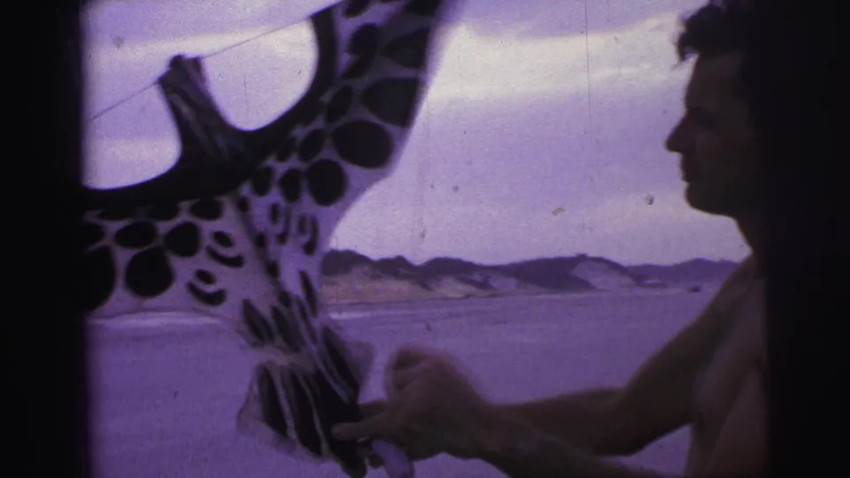 A shirtless man in partial profile stands beside the spotted kite on a Brazilian beach in 1967 home-movie footage.