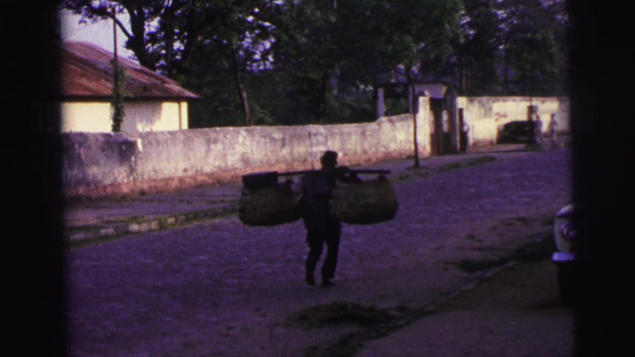 In 1967 Brazil home-movie footage, a man carries two large baskets on a pole along a cobblestone residential street.