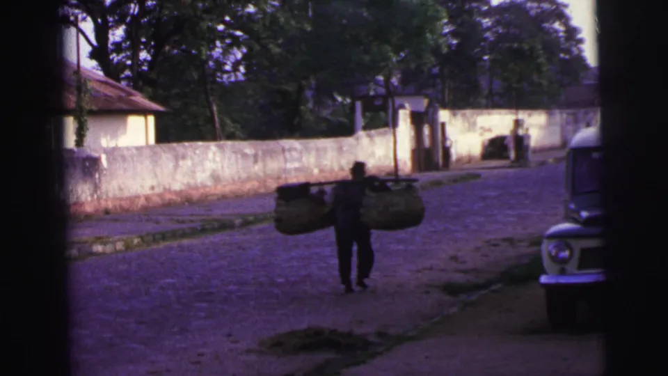 The distant basket carrier moves along a Brazilian residential road, with a weathered wall and a 1960s-era vehicle at the edge of frame.