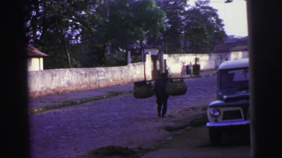 Near the end of the 1967 Brazil clip, the basket carrier recedes along the cobblestone road beside the wall and vehicle.