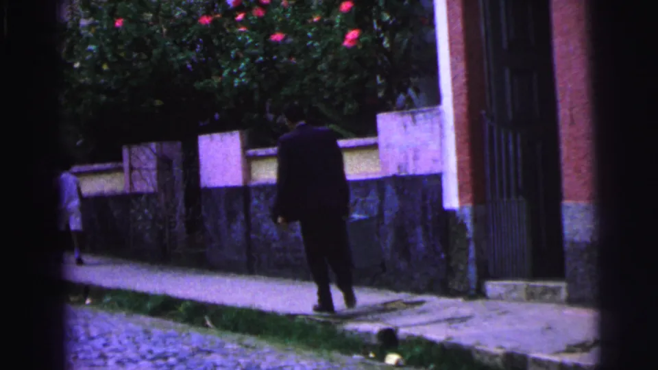 In 1967 Brazil footage, a man in a dark suit carries a briefcase along a sidewalk near a stone wall and flowering shrubs.
