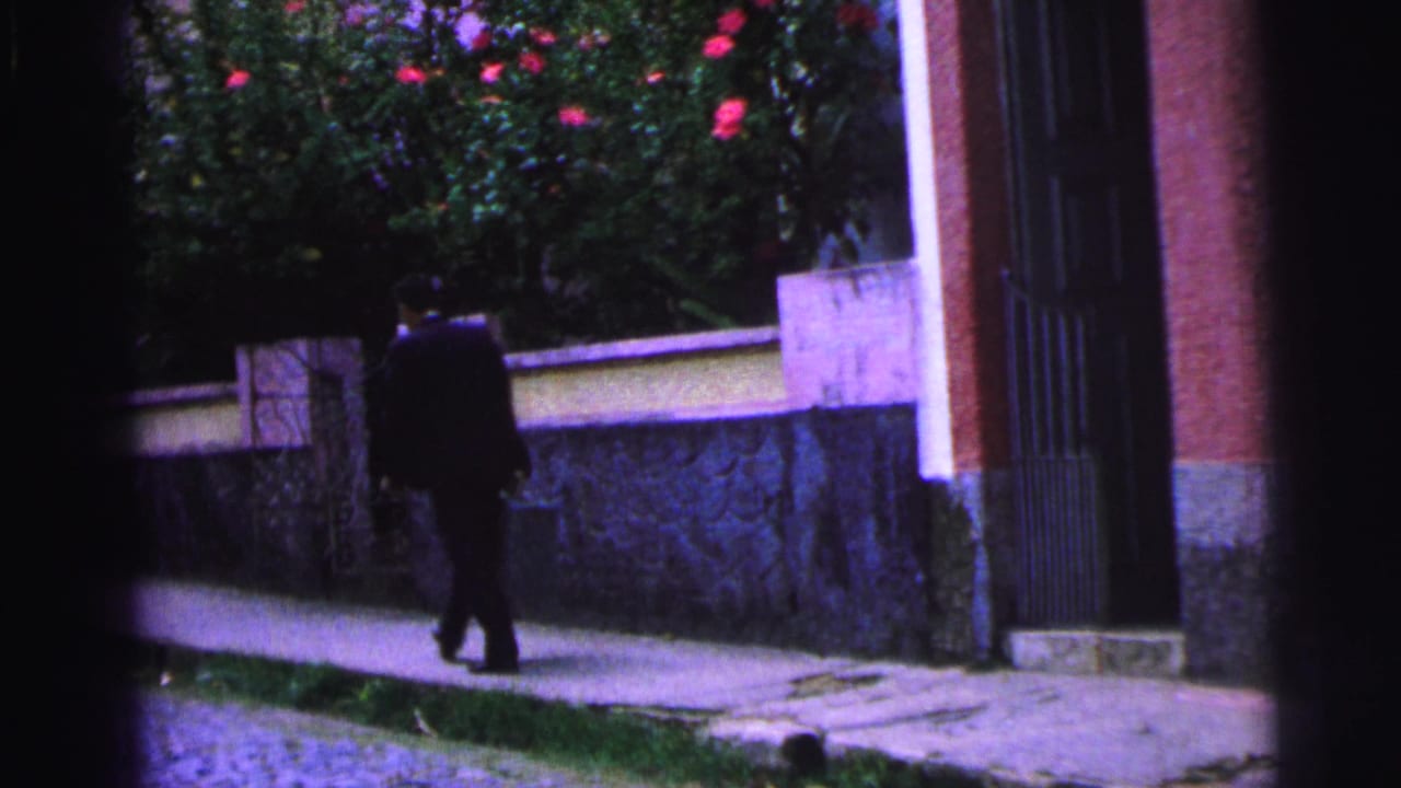 Man in a dark suit continues down the sidewalk near a gate and wall.